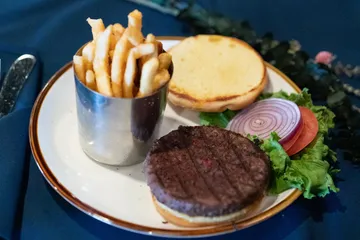 A grilled burger patty on a toasted bun with fries in a metal cup, lettuce, tomato and red onion