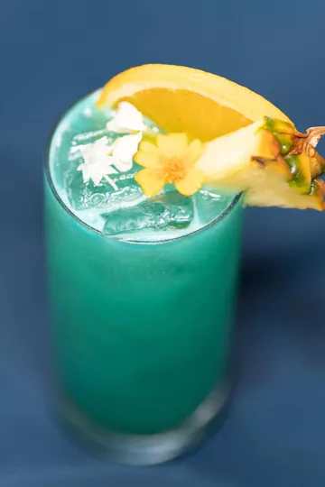 A tall turquoise cocktail over ice, garnished with an orange wheel, a pineapple wedge and edible flowers