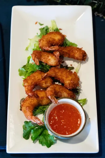 Six coconut-crusted shrimp fanned along a white plate with a ramekin of sweet chilli sauce