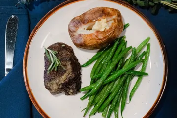 A filet mignon topped with rosemary, beside a baked potato with butter and green beans
