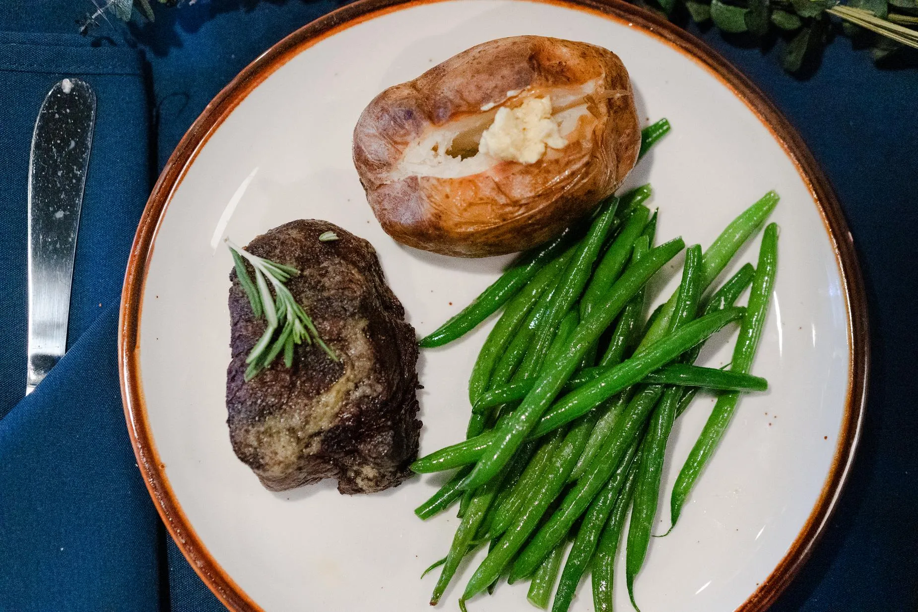 A filet mignon topped with rosemary, beside a baked potato with butter and green beans