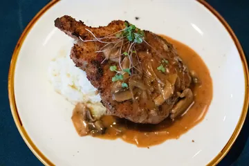 A breaded bone-in pork chop under mushroom gravy, with mashed potato and micro greens